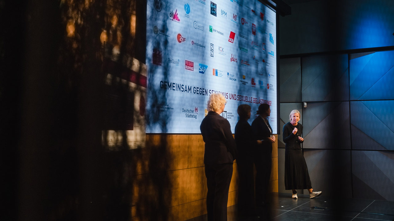 Auf dem Foto sind Claudia Hartwich (rechts) und Helene Lamprecht (links) vor den Logos der teilnehmenden Verbände, Unternehmen, zivilgesellschaftlichen Organisationen und kommunale Vertreter*innen zu sehen