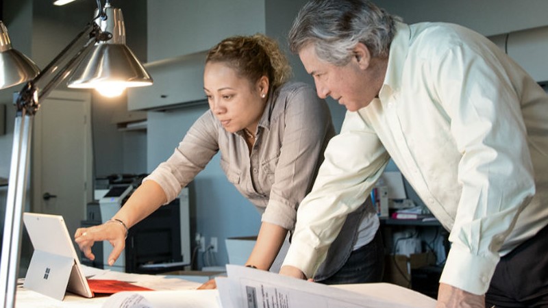 Zwei Mitarbeiter, eine Frau und ein Mann, im Gespräch an einem Tisch mit Architektur-Plänen und einem Laptop.