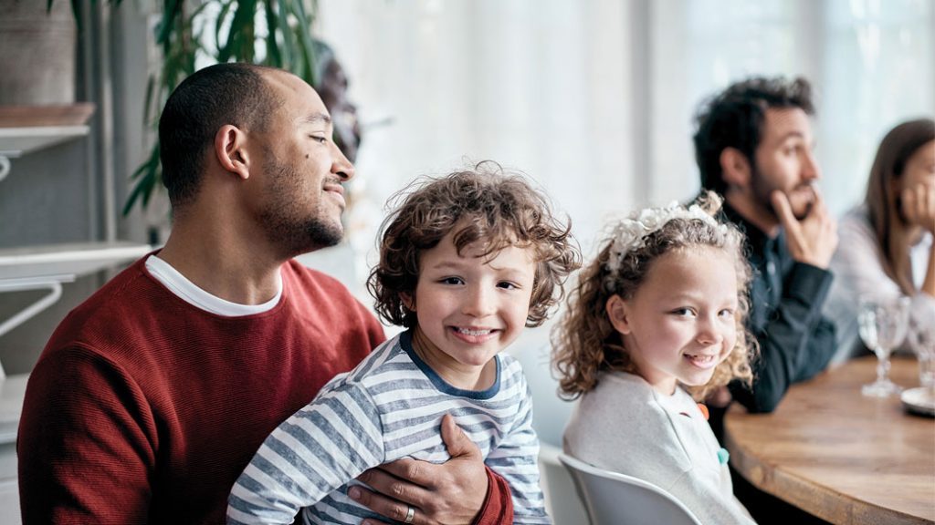 Eine Familie sitzt am Tisch. Der kleine Junge sitzt auf dem Schoß des Vaters und lächelt in die Kamera