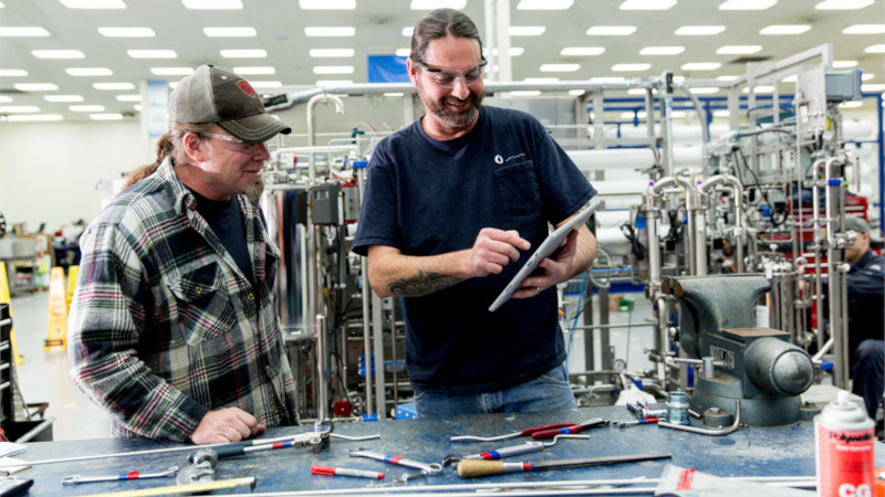 Auf dem Bild sind zwei Männer in einer Werkstatt zu sehen. Ein Mann hält ein Surface in der Hand.