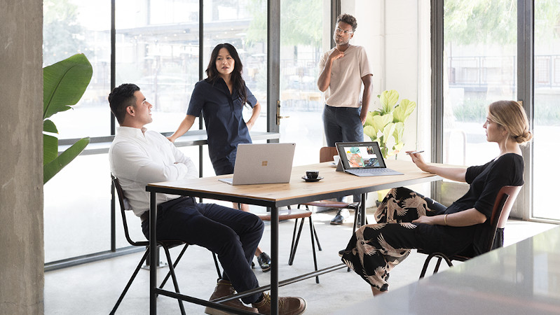 Zwei Frauen und zwei Männer sitzen bzw. stehen an einem Tisch in einem Besprechungsraum mit Surface-Geräten vor ihnen.