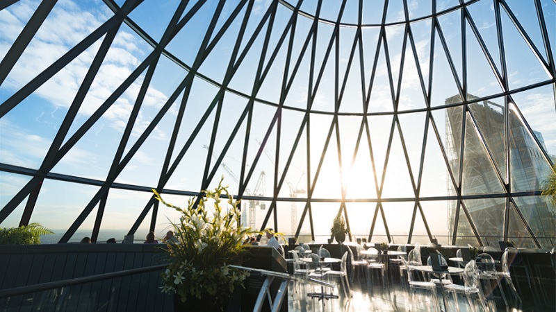 Innenansicht des Restaurants und der Cocktailbar in der obersten Etage im Londoner Gherkin-Gebäude. Restaurantgäste sitzen in klaren Acrylstühlen und Tischen. Blauer Himmel und Wolken, Baukräne und ein Wolkenkratzer sind durch die vergitterten Glasfenster der Gebäudekuppel sichtbar.