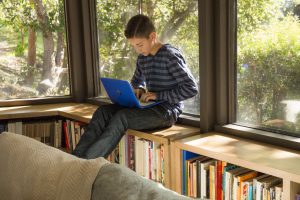 Ein Junge sitzt auf einem Bücherregal am Fenster und arbeitet an einem Laptop. Ein Junge sitzt auf einem Bücherregal am Fenster und arbeitet an einem Laptop.
