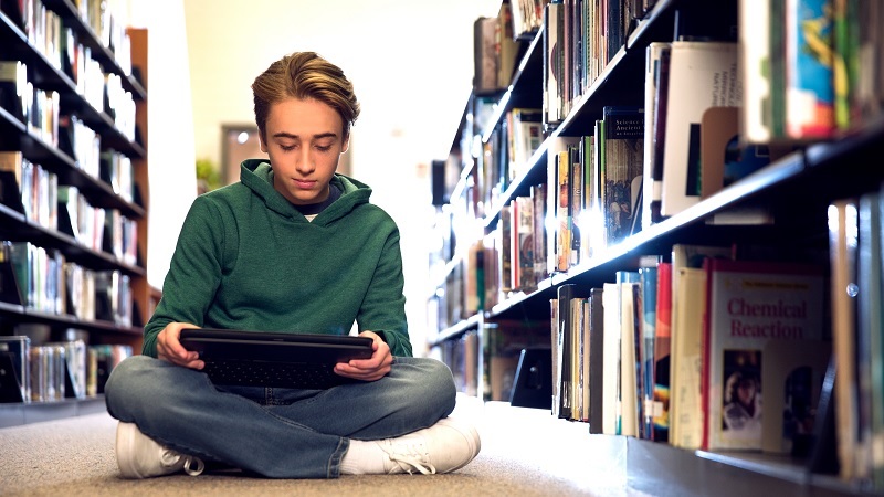 Ein Sch&uuml;ler sitzt auf dem Boden einer Bibliothek und liest ein Buch