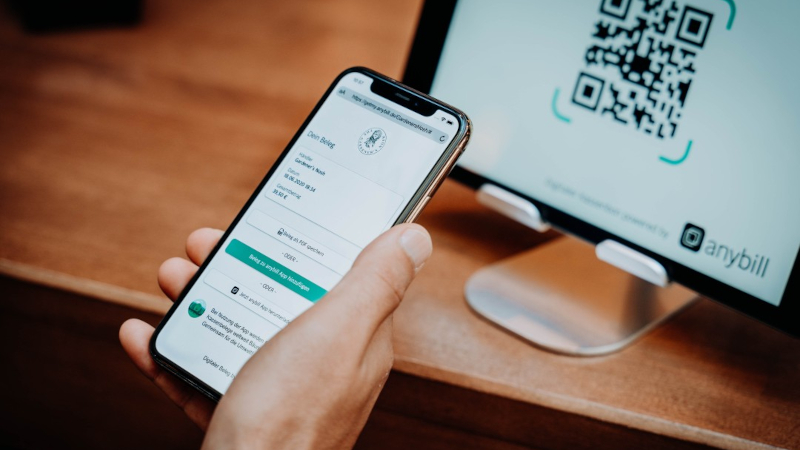 Auf dem Bildschirm eines Smartphones sieht man einen Online-Beleg. Im Hintergrund sieht man einen QR-Code, der zu dem Beleg f&uuml;hrt, auf einem Tablet