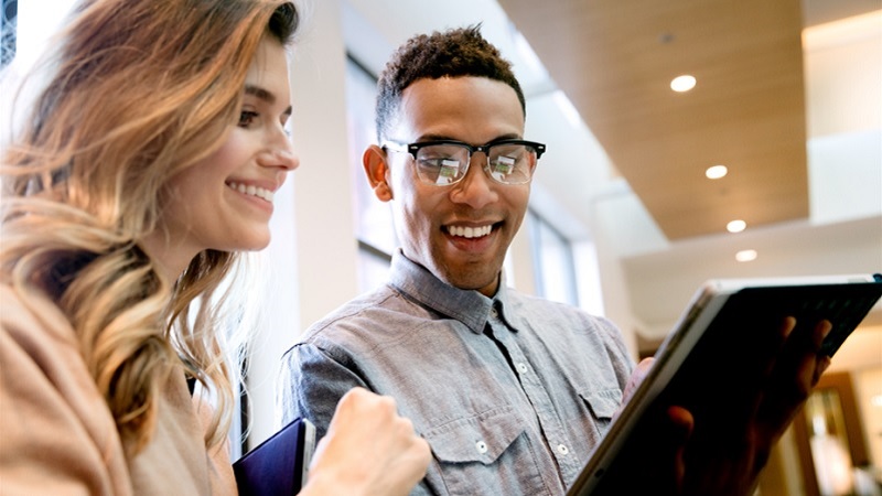Eine Studentin und ein Student im Campusgeb&auml;ude, l&auml;cheln und schauen zusammen auf das Tablet (Bildschirm nicht gezeigt)