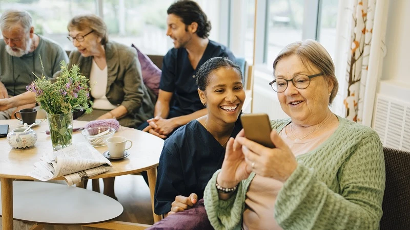 Eine &auml;ltere Dame in einer Altenpflegeeinrichtung schaut auf ihr Handy. Neben ihr kniet eine Altenpflegerin und schaut ihr dabei zu