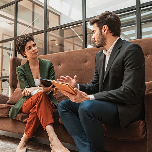 Two people dressed in business attire seated on a sofa, engaged in conversation. Two people dressed in business attire seated on a sofa, engaged in conversation.