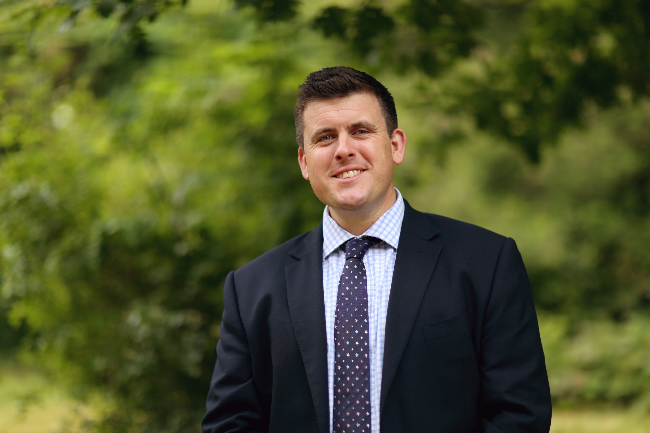 James Featherstone, a man wearing a suit and tie smiling at the camera. He is outside in front of some green bushes.