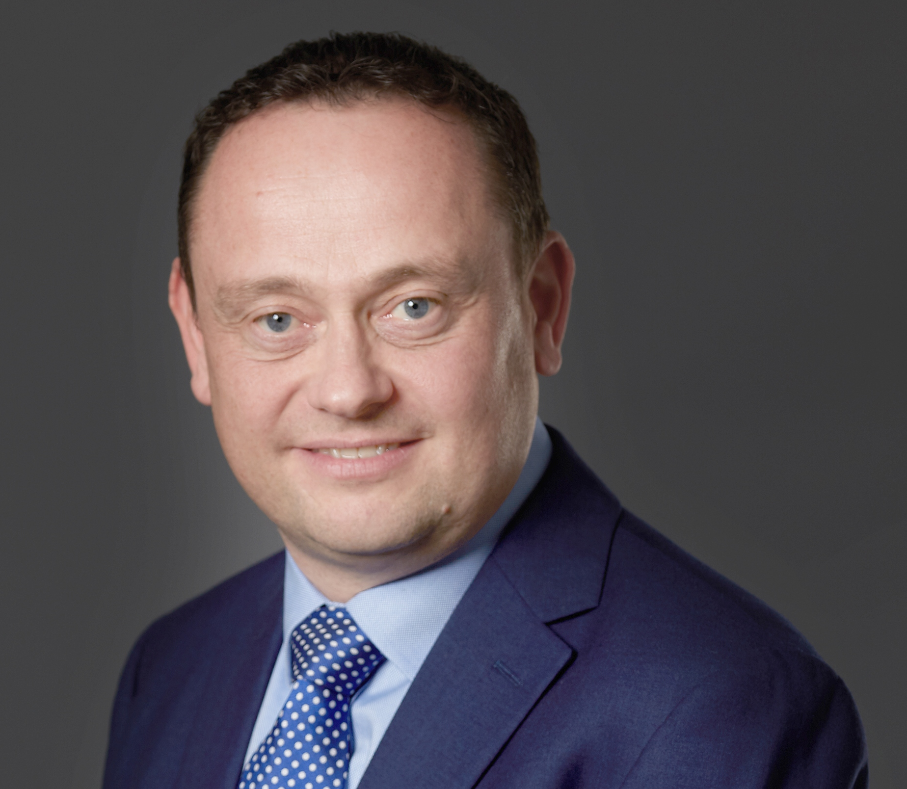 Photo of Phil Hadfield, a man with dark hair in a navy business suit with a light blue shirt and blue tie smiling at the camera