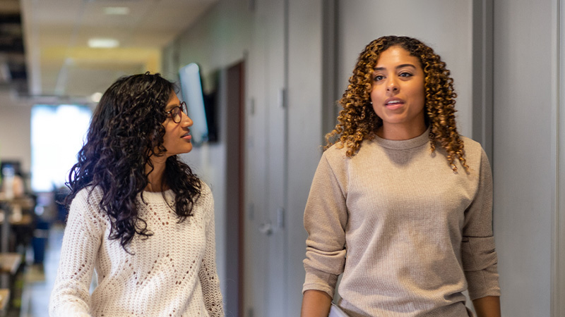 Two women walking together in an office hallway, talking as they walk