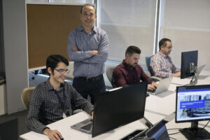 Man standing with his arms folded in front of him and smiling while three others sit before him in an office working on laptop computers.