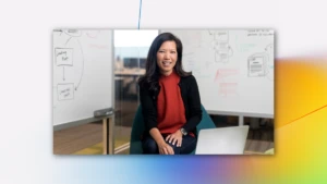 Portrait of female executive in red shirt, smiling and facing camera. She is in an office conference room setting, using an open laptop (screen not shown). Two whiteboards are partially visible in the background, with diagrams and flowcharts written on them.
