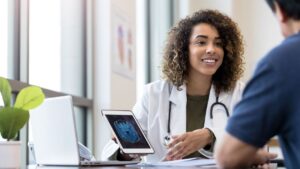 A woman in a white coat holding a tablet talking to a man