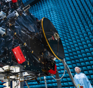 A large rectangular structure with many attachments and complex parts suspended in an enormous hangar; a woman in a sterile robe and hair cover stands on the ground at lower right.