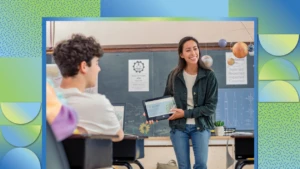 A teacher holding a folded laptop in front of students in a classroom.