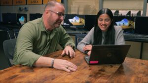 An educator and student sit together at a table in a classroom and look at the screen of a laptop.