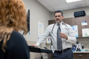 A doctor wearing a stethoscope and ID badge speaks to a patient in a medical exam room