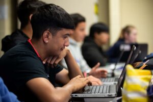 A classroom scene showing a row of students using laptops, focused on single student in the foreground.
