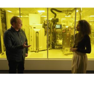 A man and woman stand in front of a window to a clean room.