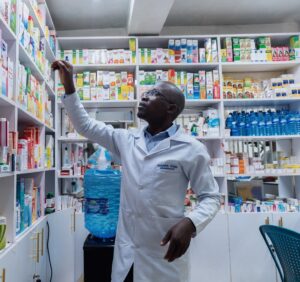A man with glasses and a white coat reaches for a high shelf of medicines.