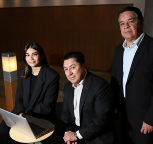 One woman and two men in a hallway, sitting on a chair with a laptop.