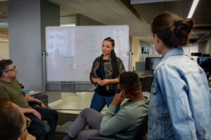 Team members gathered in an open office, listening to a colleague presenting ideas on a whiteboard.