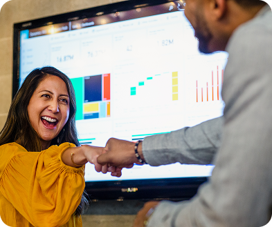 A man and woman shake hands in front of a large screen displaying a presentation or graphic.