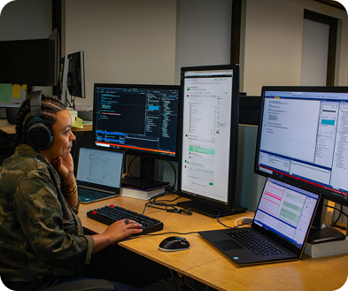 A woman seated at a desk, focused on three computer monitors displaying various applications and data.