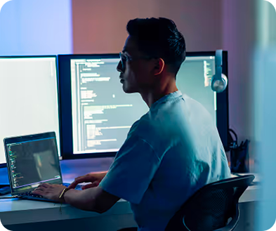 A man seated at a desk, focused on two computer screens displaying various applications and data.