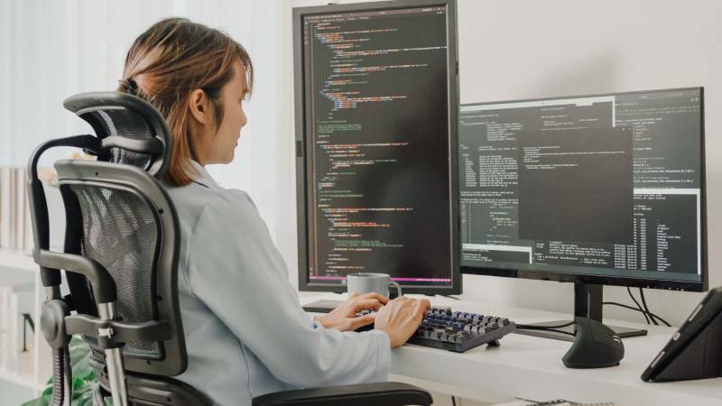 A woman seated at a desk, working on two monitors with a keyboard in front of her.
