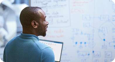 A man stands in front of a whiteboard, analyzing notes and diagrams with a focused expression.