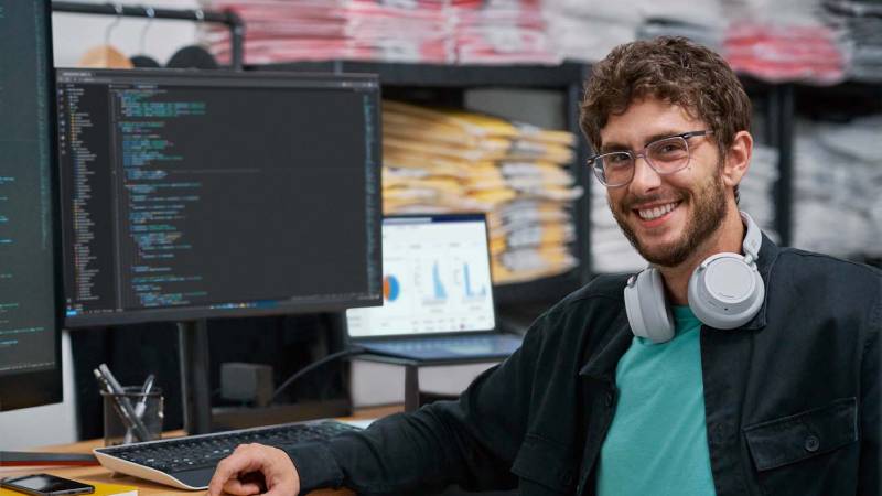 A smiling man sits in front of a computer, appearing engaged and content in his workspace.