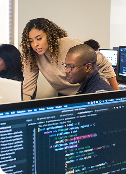 Two individuals collaborating at desks, focused on computer screens displaying lines of code.