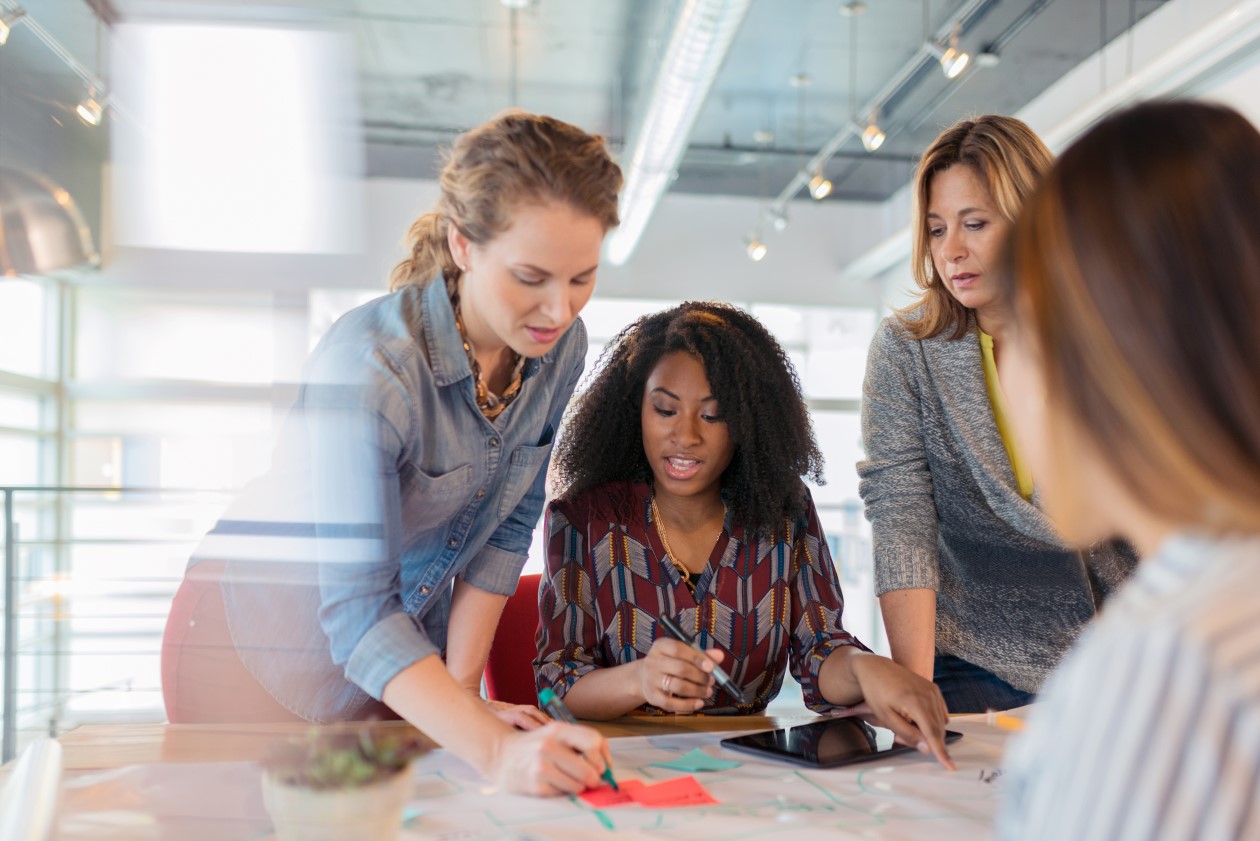 A diverse group of businesswomen look at a digital tablet and discuss a project in their modern office