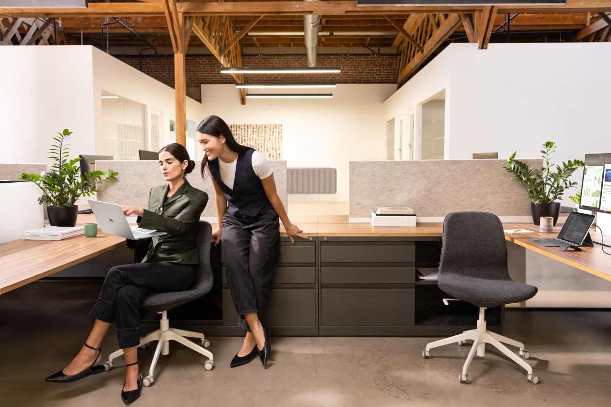 Photograph of two female executives in an office. One executive is seated, holding a Surface Laptop 13 Platinum device in her right hand. A Surface Pro 12 Platinum device in laptop mode is on another desk, connected to an external monitor. A Surface Arc Mouse is next to the device