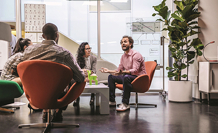A team meets in a conference room. All workers are turned toward one employee who is giving an explanation