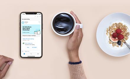 Cortana Daily Briefings on a mobile device. User holds a cup of coffee. Next to the coffee is a breakfast dish
