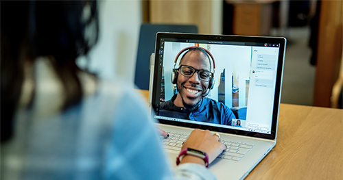 Image of a woman at a desk using a Surface laptop to make a Microsoft Teams video call with one man smiling and wearing a headset
