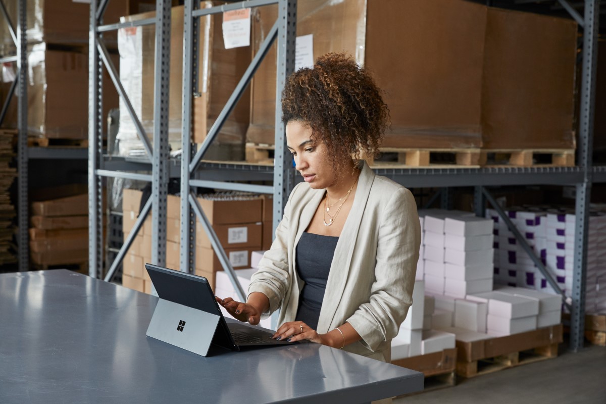 Adult female in a warehouse setting, sitting at a table using a platinum Microsoft Surface Pro X in laptop mode