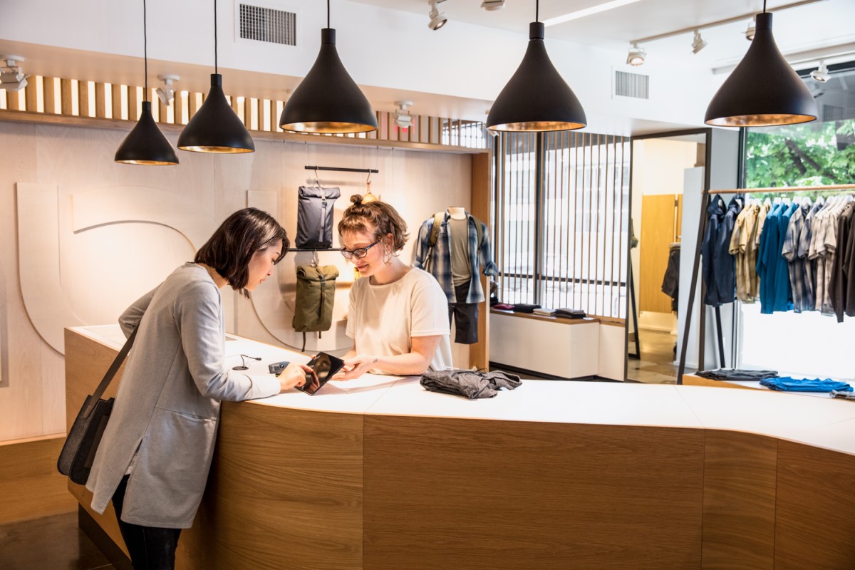 Female employee holding tablet for female consumer to sign and approve purchase. They are standing on either side of a retail clothing store checkout counter