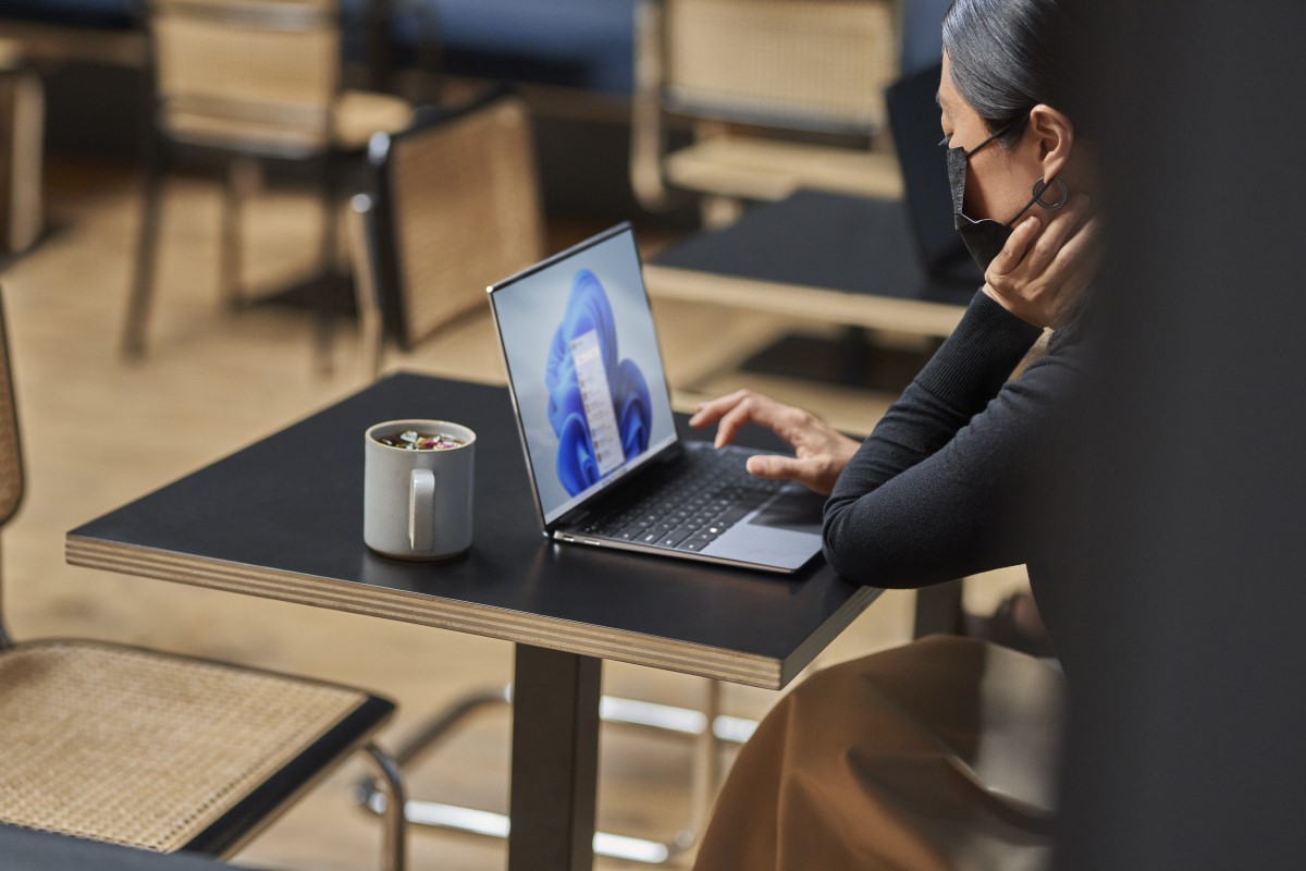 Woman with mask on for COVID compliance sitting in work space working on laptop with Windows 11 start screen image