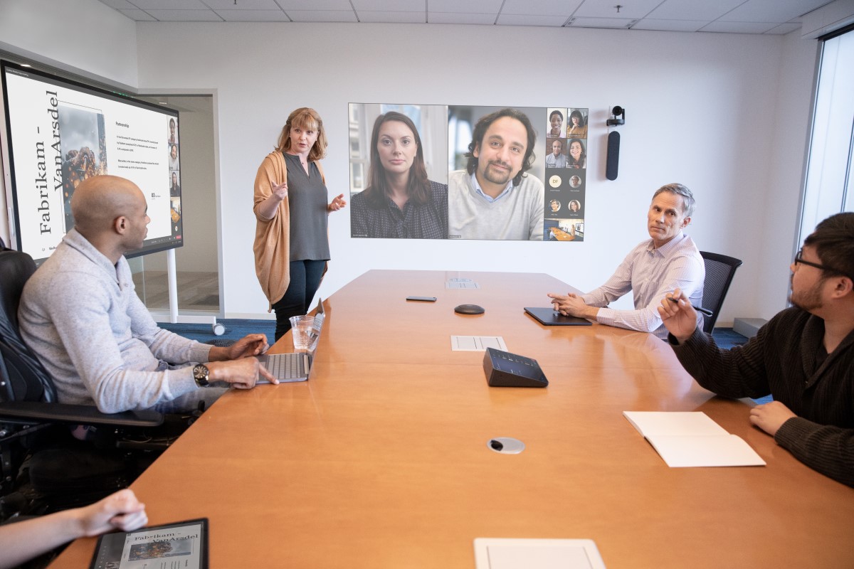 Two woman and three men (one in a wheelchair) are hosting a coordinated meeting in a large conference room and are joined by several virtual attendees on a projection screen in Front of Room view. The in-person team is interacting with remote attendees through chat and live reactions while following the woman at the Surface Hub. One woman is on an iPad logged into the Teams meeting, two men joining the meeting from an iPad and Surface