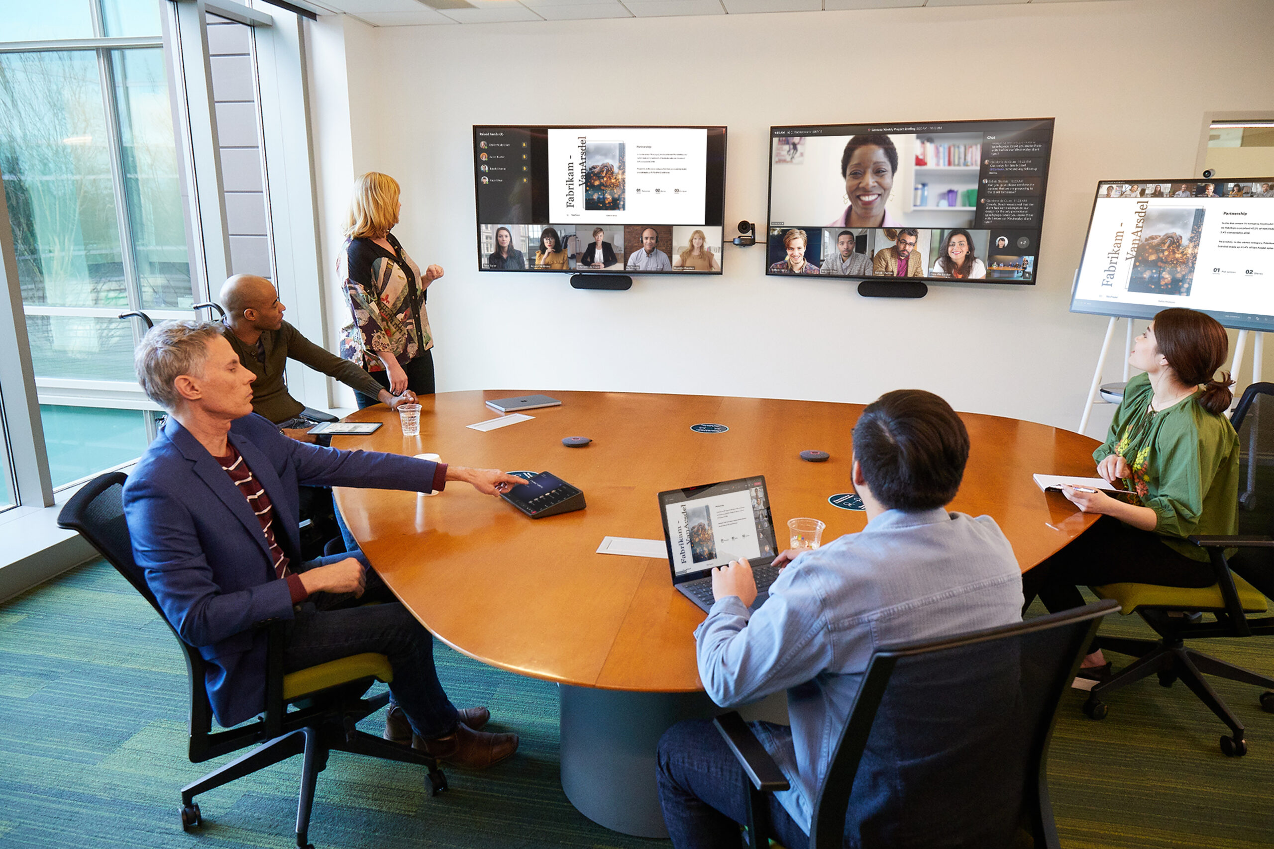 A coordinated meeting is taking place in a Microsoft Teams Room; two women, three men (one man in a wheelchair) are meeting with several team members joining remotely in Front of Room view. The in-person team is sharing a PowerPoint presentation from a Surface laptop, casting onto a Surface Hub and the Teams Room, allowing more opportunity for collaboration