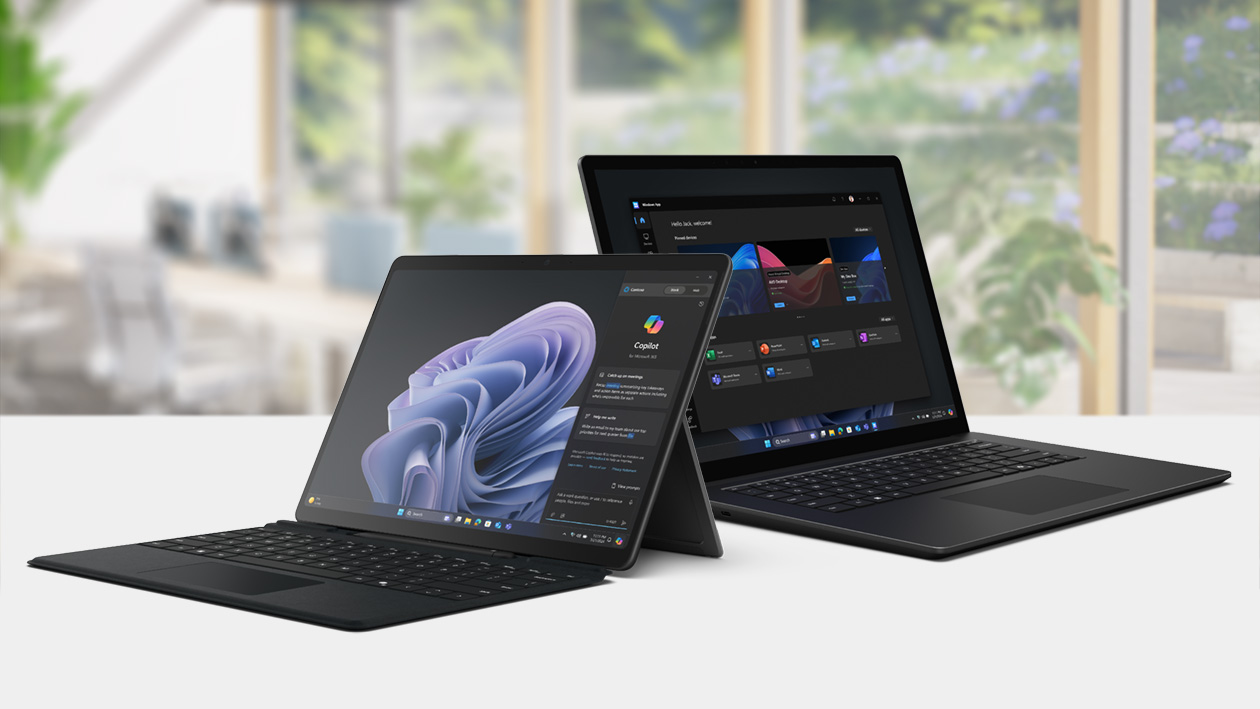 Two Microsoft Surface computer devices sitting on a table with Windows and Copilot experiences on their screens. Behind the table is an office setting in the background with tables, chairs, and windows showing a garden