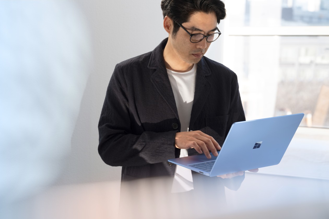 A man in a black jacket, standing and working on a laptop that hes holding