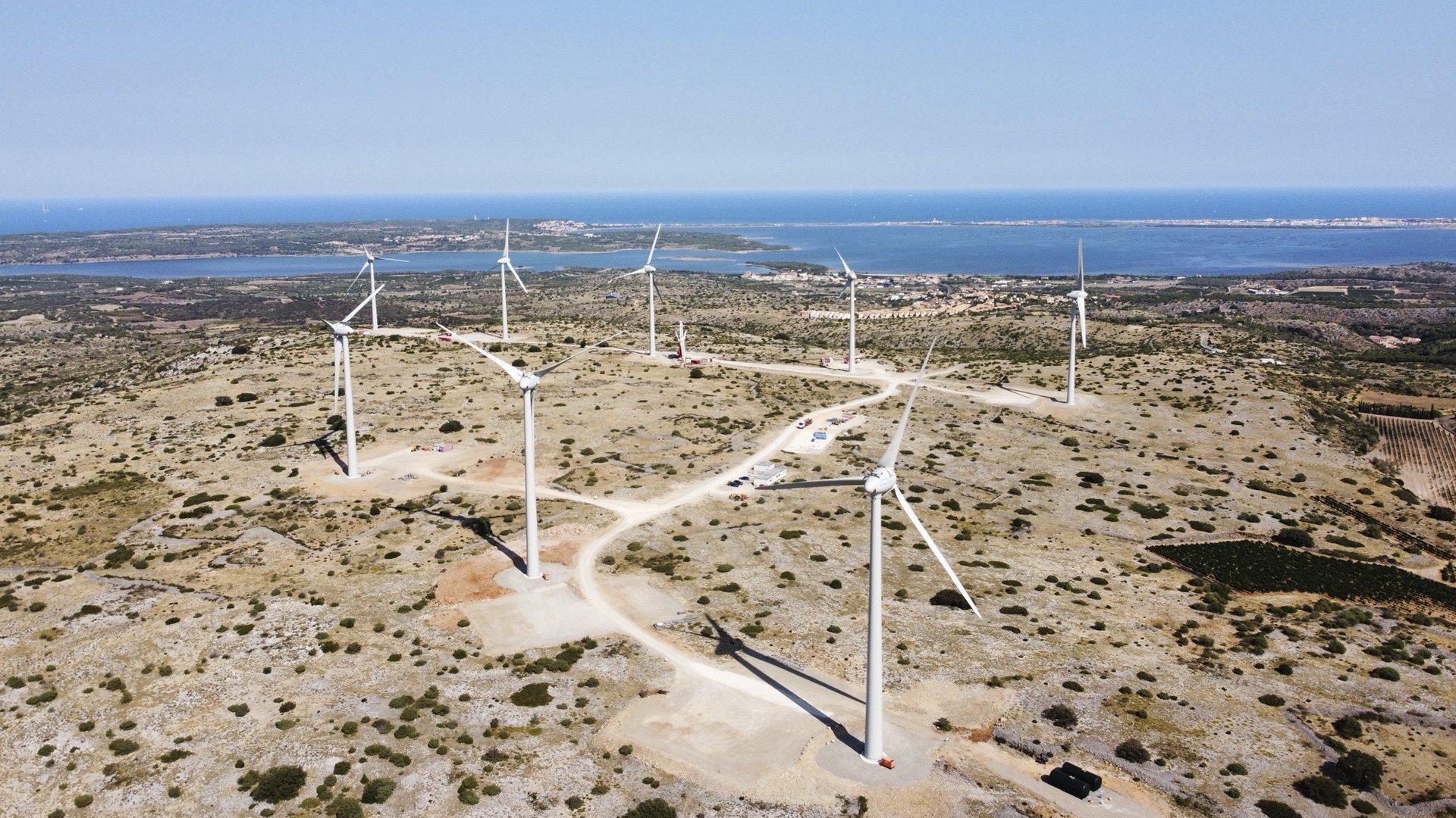Eight wind turbines top an arid hill, with the Mediterranean Sea in the distance.