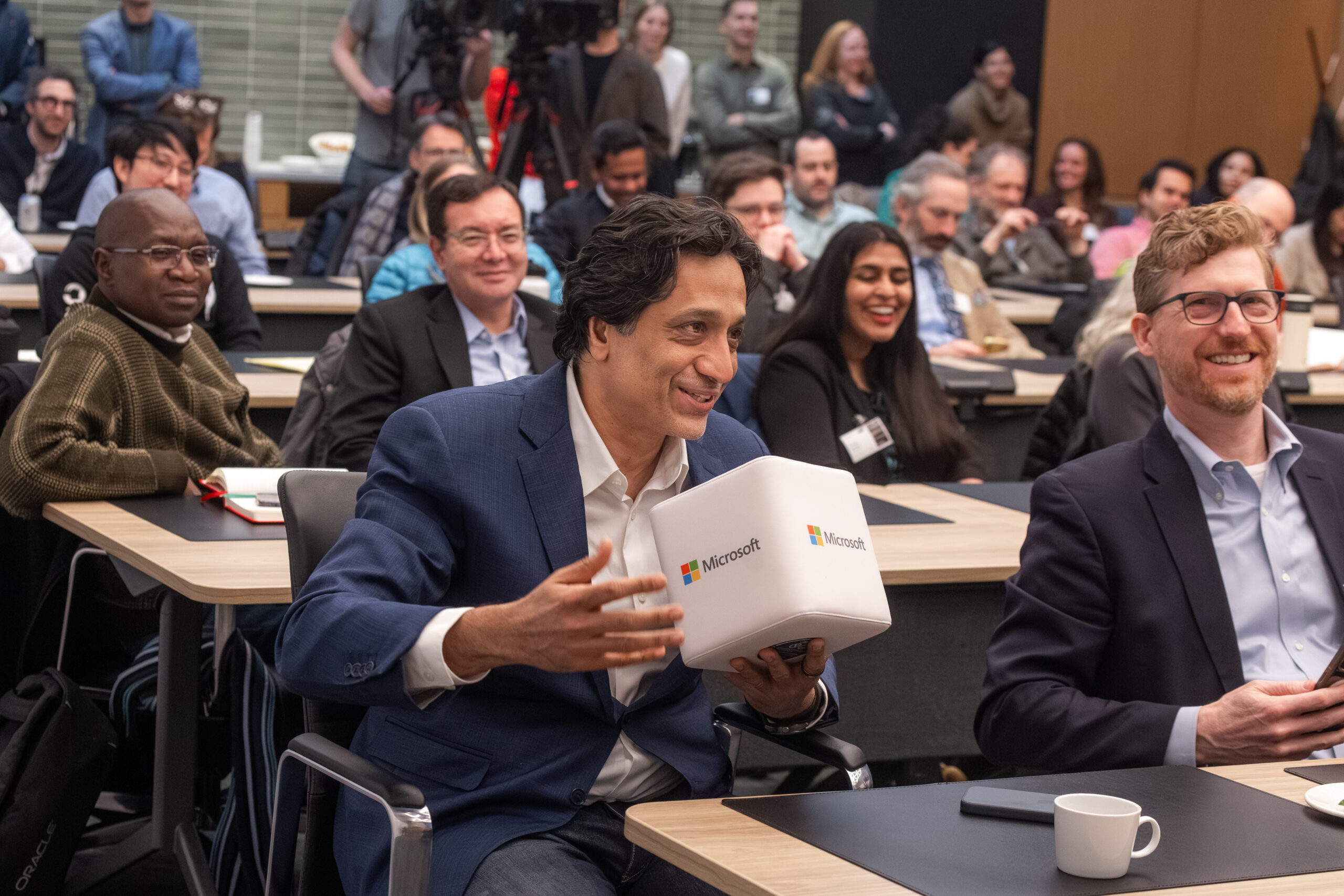 Audience members at a Microsoft AI Economy Institute event sit in a classroom-style setting, smiling and engaging as Juan Lavista Ferres in the front row holds a small Microsoft-branded device while speaking, with others listening and cameras recording in the background.