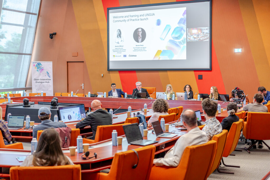 Attendees seated in a large conference room during a panel discussion, with presentation slides displayed on a screen at the front of the room.