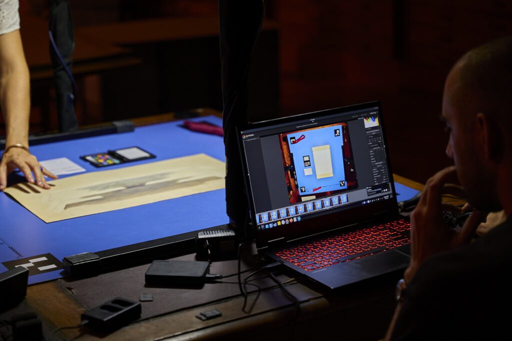 Person reviewing and editing a digitized document on a laptop while a photographed artwork or archival item is positioned on a capture table.